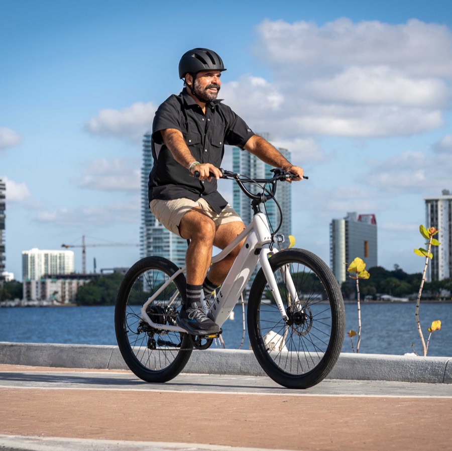 A rider on a white Pure Cycles Flux e-bike on a waterfront path, Miami skyline behind.
